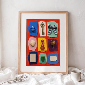 May include: A framed wall art piece featuring a bright red background with nine distinct colorful paper cutouts. The cutouts display a blue marble, a metal key, a blue and white striped bow, a seashell, a red chess pawn, a dried poppy pod, a beige rectangle, a green cube, and a blank white label.