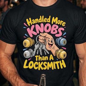 Può includere: Una t-shirt nera a maniche corte con una stampa grafica che riporta il testo Handled More KNOBS Than A LOCKSMITH. Il design include una mano in stile cartoon che tiene delle chiavi circondata da quattro pomelli colorati.