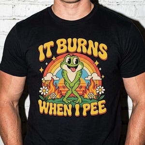 May include: A black short-sleeve t-shirt featuring a graphic of a green frog standing in front of a rainbow and flames. The text "IT BURNS WHEN I PEE" is printed in yellow, arched letters above and below the frog illustration on the chest.