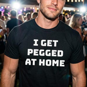 May include: A black short-sleeved crew neck t-shirt featuring the white text I GET PEGGED AT HOME printed in a bold, sans-serif font across the chest. The garment is made of a solid black fabric with a standard fit and a simple round neckline.