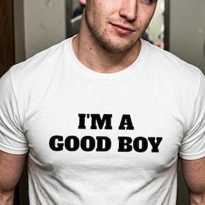 Pode incluir: Uma t-shirt branca de manga curta com o texto "I'M A GOOD BOY" impresso em letras maiúsculas pretas e em negrito no peito. A peça tem um decote redondo clássico e um corte padrão, feita de tecido branco liso.