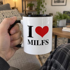 Puede incluir: Una taza de cerámica blanca con asa y borde interior negros. La parte frontal de la taza presenta el texto negro "I" seguido de un símbolo de corazón rojo y el texto negro "MILFS" impreso en una fuente sans-serif negrita sobre el fondo blanco.
