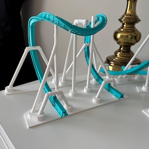 Puede incluir: Una maqueta de montaña rusa con una pista de color turquesa brillante sostenida por una estructura blanca. El modelo está montado sobre una base blanca plana y muestra una sección de pista curva con varios pilares de soporte y vigas.
