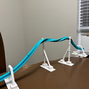 May include: A miniature roller coaster model featuring a bright turquoise track supported by three white vertical pillars with rectangular bases. The track curves in a wave-like motion, elevated above the surface by the white support structures.