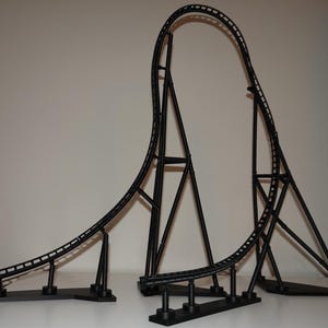 Puede incluir: Un modelo de montaña rusa en color negro que presenta una subida vertical pronunciada y una sección de caída curva. La estructura está sostenida por pilares negros delgados y descansa sobre bases planas, mostrando un diseño de riel detallado.