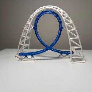 Puede incluir: Un modelo impreso en 3D de un bucle de montaña rusa con una vía azul sostenida por una estructura de arco de celosía blanca. La vía forma un bucle vertical continuo y todo el conjunto está montado sobre pequeñas placas base rectangulares.