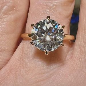 Könnte beinhalten: Ein Goldring mit einem großen, runden, im Brillantschliff geschliffenen, klaren Edelstein, der von sechs Krappen gehalten wird. Der Stein weist zahlreiche Facetten auf, die das Licht reflektieren und für ein helles, funkelndes Aussehen auf dem glatten, polierten Goldring sorgen.