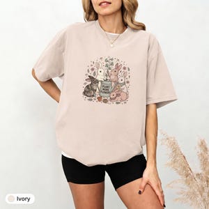 May include: An ivory short-sleeved t-shirt featuring a central graphic of five rabbits gardening. The illustration includes a watering can with the text "PLANT MORE FLOWERS" surrounded by small flowers, leaves, and ferns in muted earth tones.