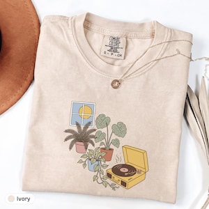 Könnte beinhalten: Ein elfenbeinfarbenes Kurzarm-T-Shirt mit einem Comfort Colors-Etikett, das einen Grafikdruck eines Fensters mit gelber Sonne, drei Topfpflanzen und einem gelben Plattenspieler zeigt. Eine dünne silberne Kette liegt am Halsausschnitt.