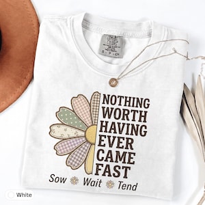 Peut inclure: Un t-shirt blanc à manches courtes orné d'un motif de demi-marguerite aux pétales à motifs dans des tons doux. Le texte indique "NOTHING WORTH HAVING EVER CAME FAST" suivi de "Sow Wait Tend" avec de petites icônes de marguerites.