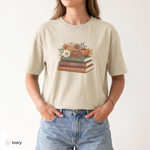 Maglietta personalizzata con pila di libri, nome personalizzato sul dorso, motivo floreale autunnale con zucche, colori rilassanti, regalo per gli amanti dei libri.