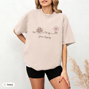 May include: An ivory short-sleeved t-shirt featuring a graphic print of a tangled line that transitions into a blooming pink flower. Below the illustration, the text "Grow Anyway." is printed in a dark brown, cursive-style font.