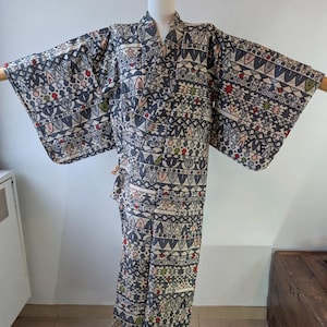 May include: A full-length kimono featuring a repeating geometric pattern in shades of charcoal gray, cream, and black. The fabric is decorated with small, stylized motifs in muted red, green, and blue, arranged in horizontal bands across the garment.
