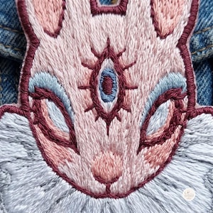 May include: An embroidered patch featuring a stylized rabbit face in shades of pale pink, light blue, and dark maroon. The design includes a central third eye detail with radiating lines, stitched onto a blue denim fabric background.