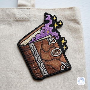May include: An embroidered patch featuring a brown book with a stitched cover, a central eye detail, and silver snake accents. Purple magical smoke rises from the open pages, accented by small yellow stars, all outlined in a thick black border.