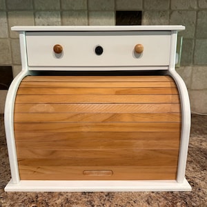 May include: A white wooden bread box featuring a roll-top door made of horizontal light brown wooden slats. The unit includes a top drawer with two small round wooden knobs and a central black circular accent piece on the front panel.