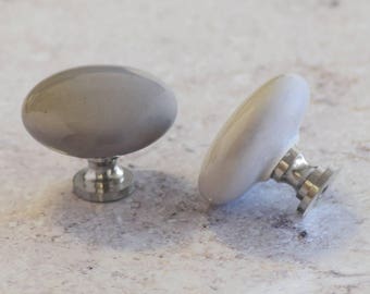 May include: Two oval-shaped cabinet knobs with a smooth, glossy gray finish. Each knob features a polished silver-toned metal base and a rounded, solid-colored ceramic top, designed for attachment to furniture or cabinetry doors.