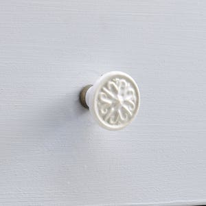 May include: A round white ceramic drawer knob with a raised, intricate, symmetrical scrollwork pattern on its face. The knob is attached to a light gray painted wooden drawer front by a small, metallic, cylindrical base piece.