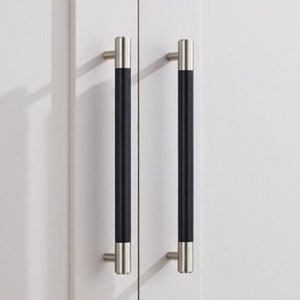 May include: Two cabinet pulls with a black centre and silver-toned ends are mounted on a white cabinet door. The pulls are vertically oriented, providing a sleek, modern aesthetic. The hardware is designed for easy gripping and opening.