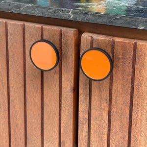 May include: Two circular cabinet knobs with bright orange centers and black rims, mounted on vertical-slatted brown wooden cabinet doors. The knobs feature a flat, smooth surface and a simple, clean design that contrasts with the wood grain.
