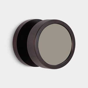 Puede incluir: Un espejo de pared redondo de color bronce oscuro, con un marco exterior estriado. El espejo tiene una superficie reflectante gris y un dise&ntilde;o simple y moderno. El di&aacute;metro es de aproximadamente 20 cm, adecuado para varios estilos de interior.