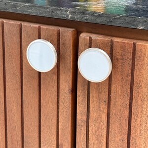 以下が含まれることがあります： メタリックシルバーの縁取りが施された2つの円形で白いキャビネットノブが、縦に溝が入った茶色の木製キャビネットの扉に取り付けられています。ノブは滑らかで平らな表面をしており、質感のある木製パネルの上に並んで配置されています。