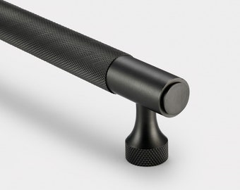 May include: A matte black cabinet pull featuring a cylindrical bar with a textured diamond knurled grip pattern. The handle is supported by a smooth, flared post that connects to a circular base with a matching knurled edge detail.