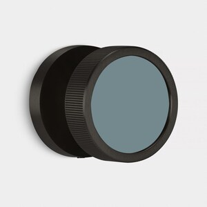 May include: A black, circular wall mirror with a ridged edge and a muted teal-colored glass. The mirror is mounted on a white wall, showcasing a minimalist design. The mirror's diameter is approximately 6 inches.