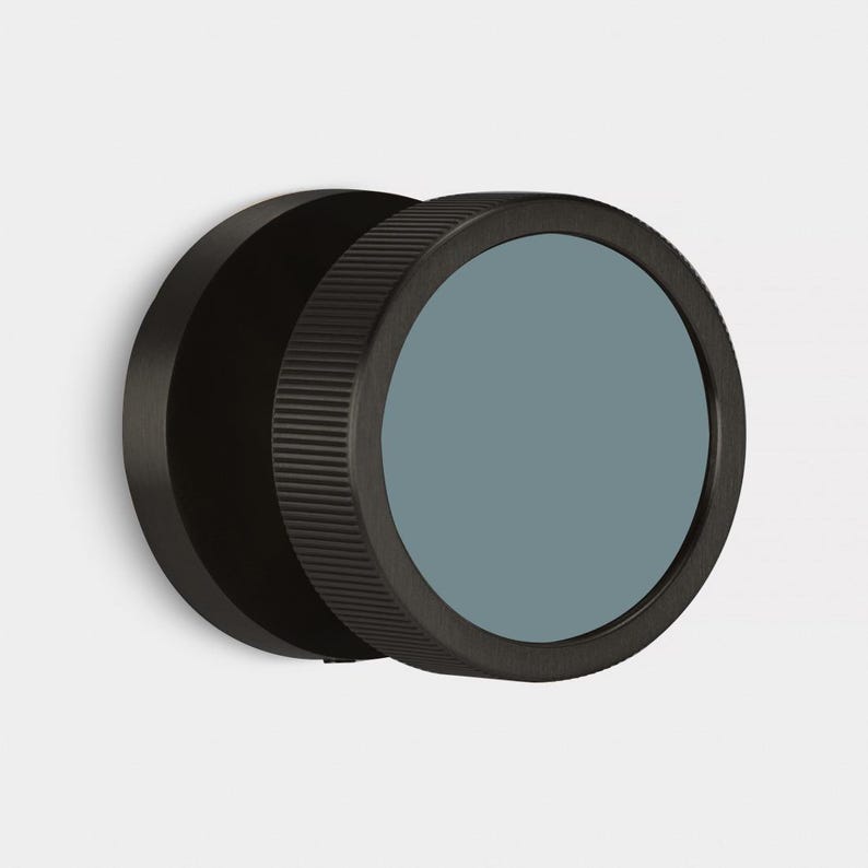 May include: A black, circular wall mirror with a ridged edge and a muted teal-colored glass. The mirror is mounted on a white wall, showcasing a minimalist design. The mirror's diameter is approximately 6 inches.