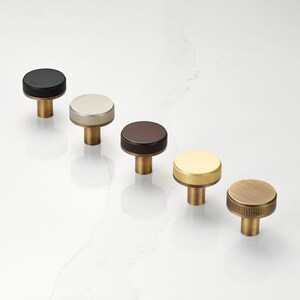 May include: Five cabinet knobs in different finishes. Each knob features a circular, ridged top and a cylindrical stem. The colours are black, silver, brown, gold, and brass. These metal knobs are suitable for use on furniture.