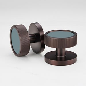 Puede incluir: Dos pomos de puerta de color bronce con un dise&ntilde;o circular. Cada pomo tiene un anillo exterior cil&iacute;ndrico estriado y una base circular plana. El centro de cada pomo presenta una incrustaci&oacute;n circular de color verde azulado. Los pomos est&aacute;n dise&ntilde;ados para una est&eacute;tica moderna.