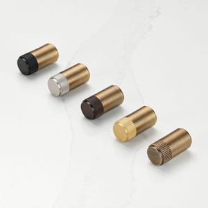 Puede incluir: Cinco tapones de botella cilíndricos con cuerpos de latón y varios acabados de tapa final: negro, plateado, marrón oscuro, dorado y bronce. Los tapones están dispuestos en diagonal sobre una superficie de mármol blanco.