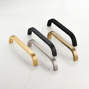 May include: An assortment of cabinet handles in various metallic finishes: gold, silver, black, and brass. The handles have a curved, rectangular design, suitable for drawers or cabinet doors. The hardware is displayed on a white surface.