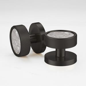 Op de afbeelding: Drie zwarte kastknoppen met een cirkelvormig ontwerp. De knoppen hebben een getextureerde, geribbelde rand en een gespikkelde, grijs en witte kern. Eén knop staat rechtop, terwijl de andere twee op hun kant liggen.