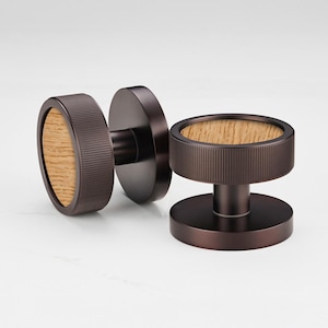 Puede incluir: Pomos de puerta de color bronce con un centro de vetas de madera. Los pomos tienen un diseño circular con un borde exterior estriado. La veta de la madera es de color marrón claro. Los pomos se muestran sobre un fondo blanco.