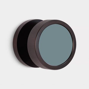 Puede incluir: Un espejo de pared redondo de color bronce oscuro, con un borde exterior estriado y un cristal verde azulado mate. El espejo est&aacute; montado en la pared, destacando su dise&ntilde;o minimalista y su est&eacute;tica moderna.