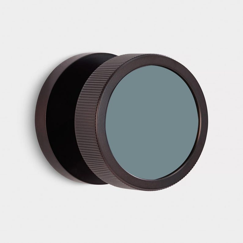 Puede incluir: Un espejo de pared redondo de color bronce oscuro, con un borde exterior estriado y un cristal verde azulado mate. El espejo est&aacute; montado en la pared, destacando su dise&ntilde;o minimalista y su est&eacute;tica moderna.