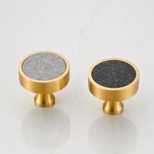 May include: Two circular cabinet knobs with a brushed gold metal finish. One knob features a light gray speckled stone inlay on the face, while the other knob features a dark charcoal gray speckled stone inlay on the face.