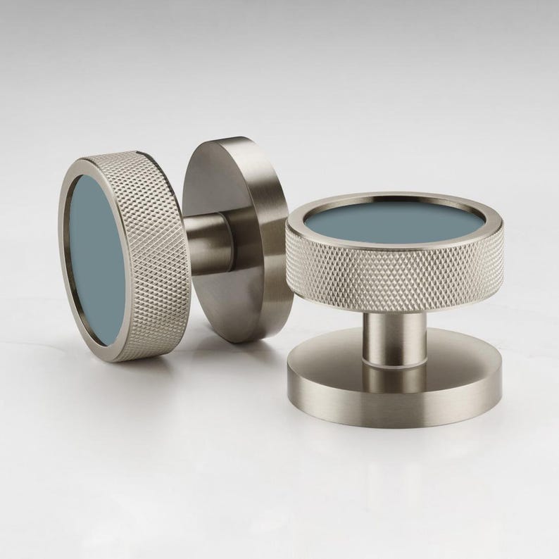 Puede incluir: Tres pomos de armario de n&iacute;quel cepillado con un anillo exterior texturizado y un centro azul claro. Los pomos tienen un dise&ntilde;o moderno y son adecuados para cajones o puertas de armarios. Los pomos se muestran sobre un fondo blanco.