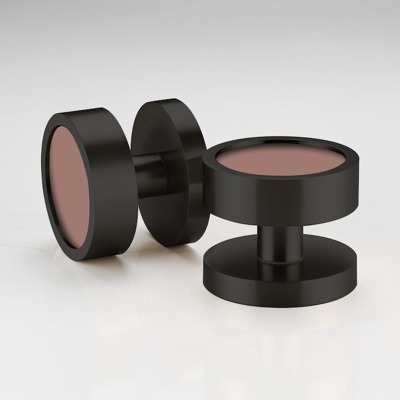 May include: Three black, cylindrical cufflinks with a circular, muted rose-colored center. The cufflinks are displayed on a white surface. The design is modern and minimalist.