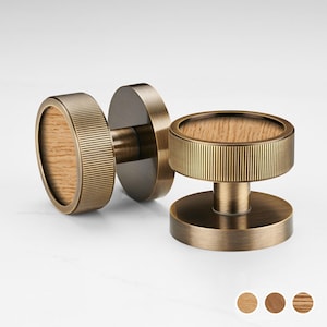 Pode incluir: Duas maçanetas de porta de latão antigo com um centro de grão de madeira. Os botões têm um design circular com uma borda externa canelada. O centro do grão de madeira é de cor castanho claro. Os botões estão em um fundo branco.