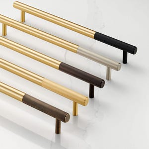 Puede incluir: Una colección de tiradores de armario dorados con extremos texturizados y coloreados. Los tiradores tienen un diseño cilíndrico con una barra lisa y recta y un agarre texturizado contrastante en negro, plata, marrón y dorado.