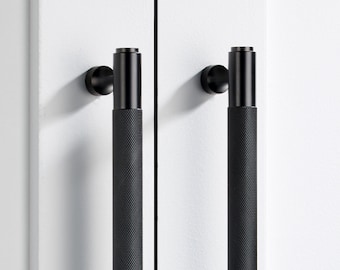 May include: Two matte black cylindrical cabinet handles with a textured knurled grip pattern. Each handle features a smooth top cap and a circular mounting base with a ridged edge, attached vertically to a white cabinet door surface.
