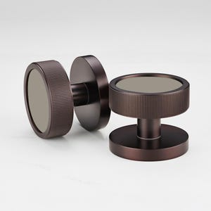 Può includere: Due pomelli per porte color bronzo con un design circolare. Ogni pomello presenta un anello esterno cilindrico scanalato e un centro piatto e circolare. I pomelli sono posti su uno sfondo bianco, mostrando la loro estetica moderna.