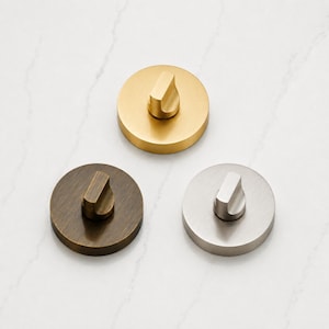 May include: Three circular door thumb turns arranged in a triangle. One is brushed gold, one is dark bronze, and one is brushed silver. Each piece features a flat, round base with a central, vertical, rectangular knob for manual operation.