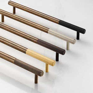 Peut inclure: Une sélection de poignées de meubles dans différentes finitions. Les poignées ont un design cylindrique avec une texture côtelée sur les extrémités des poignées. Les couleurs incluent le bronze, le noir, l'argent, le marron et l'or. Les poignées sont disposées en diagonale sur une surface blanche.