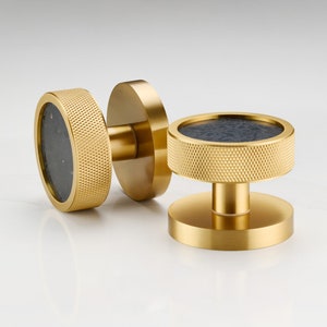 Pode incluir: Dois botões de armário dourados com um centro preto. Os botões têm um design texturizado e estriado.