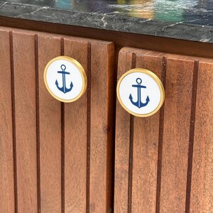Puede incluir: Dos pomos circulares para armarios con borde de metal dorado e incrustación central blanca. Cada pomo muestra un gráfico azul oscuro centrado de un ancla náutica. Los pomos están montados en puertas de armario de madera marrón con listones.