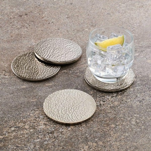 May include: A set of four circular silver-toned metal coasters with a hammered, textured surface. One coaster supports a clear glass filled with ice cubes and a yellow lime wedge, while the other three coasters are arranged nearby.