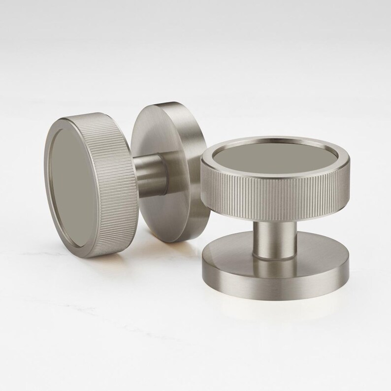 Może przedstawiać: Trzy gałki do szafek ze szczotkowanego niklu. Każda gałka ma okrągły kształt z żebrowanym brzegiem i płaskim, okrągłym środkiem. Gałki są przeznaczone do łatwego montażu i dodają nowoczesnego charakteru każdej szafce lub szufladzie.